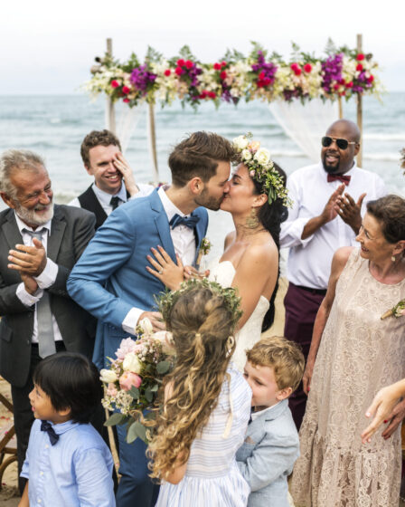 Bride and groom kissing at beach wedding ceremony, surrounded by family and floral decorations.