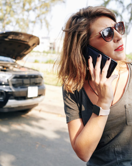 Woman on phone next to car with open hood, discussing car insurance claim.