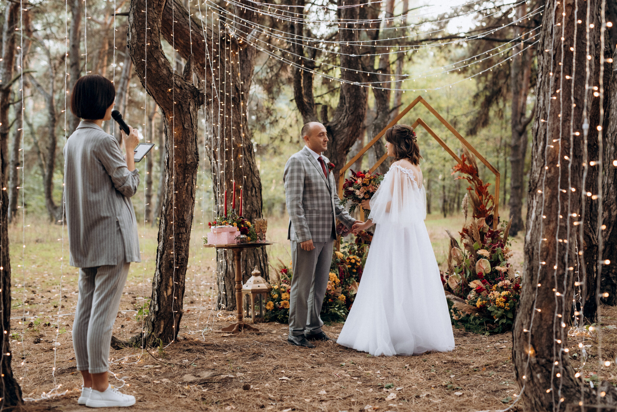 Couple exchanging vows in a forest wedding setting with floral arch and string lights.